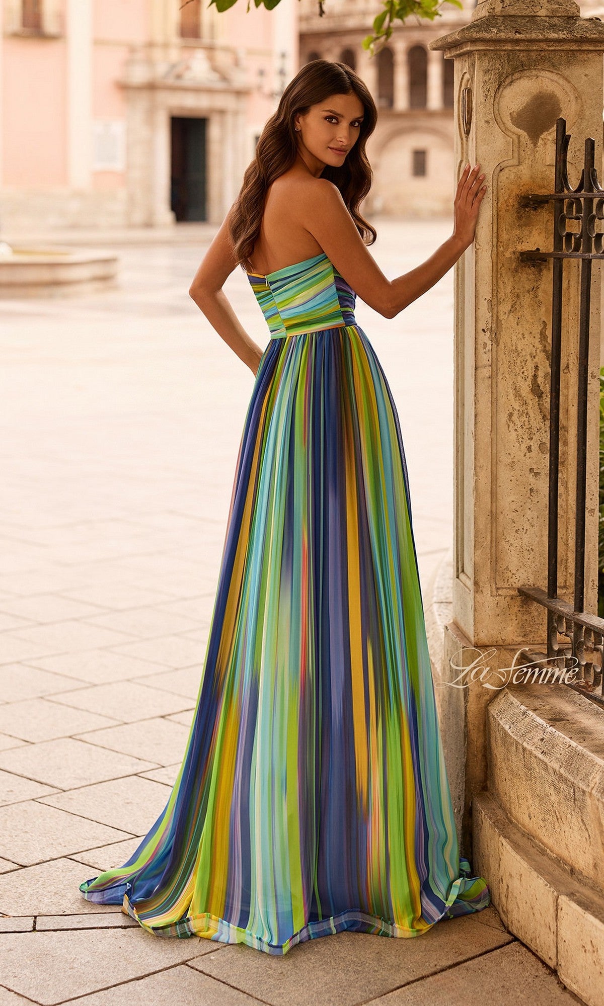 A woman wears the La Femme 33690 multi-colored prom dress, featuring vibrant vertical stripes, as she stands outdoors with one hand on a stone column, glancing over her shoulder.